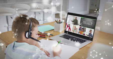 Writing boy in striped shirt with headset taking notes at home kitchen area, with laptop....