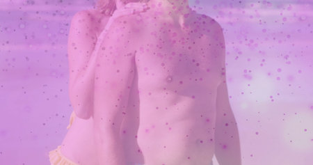 Embracing pair wearing dark swim trunks and yellow bikini bottom at beach shoreline, pink overlay....