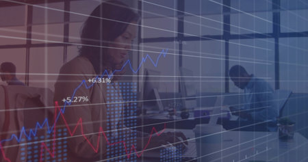 Typing woman wearing blazer working on laptop in open-plan office, with financial graphs overlay....