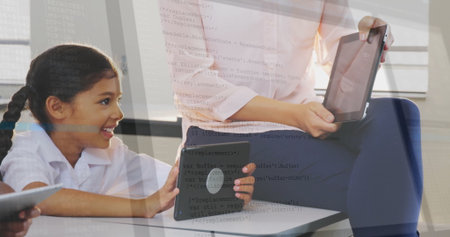 Holding tablet teacher guiding girl in collared shirt leaning at classroom desk, with second tablet....
