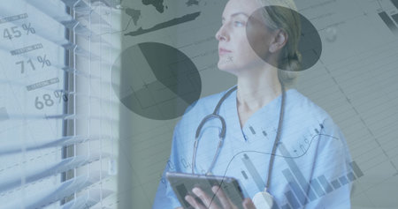 Clinician wearing scrubs with stethoscope, studying data charts on tablet by blinds in clinic....