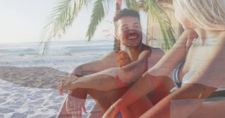 Smiling friends in swimwear relaxing on white sand beach under palm tree frond, ocean waves behind....