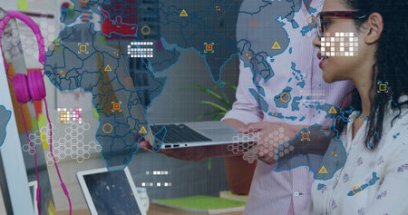 Standing colleagues wearing office attire, viewing laptop screen at office with world map overlay....