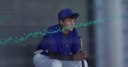 Sitting teen in blue uniform and cap gripping wooden bat on dugout bench, gazing toward field....