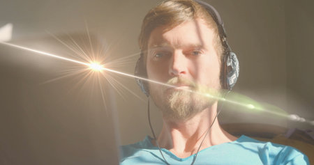 Concentrating man wearing light-blue shirt and headphones working inside room, with laptop screen....
