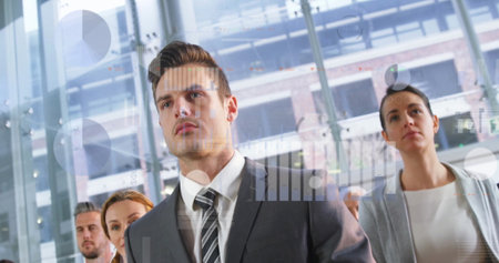 Standing business professionals displaying digital charts in glass-walled atrium with steel columns. Corporate, teamwork, analytical, futuristic, polished, innovation, leadershipの写真素材