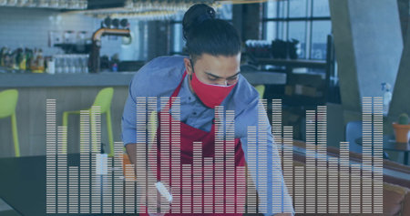 Leaning staff wearing red apron and mask wiping dining table in bar area, with spray bottle....