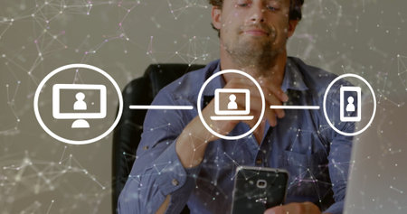 Interacting man wearing blue shirt tapping smartphone at office desk, with laptop, network icons....