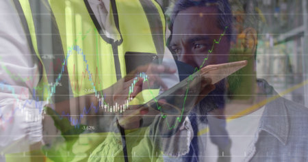 Analyzing men wearing safety vest using smartphone and tablet in warehouse, with financial chart. Industrial, teamwork, technology, analytics, collaboration, modern, logisticsの写真素材