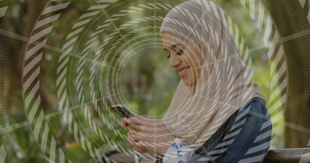 Smiling woman wearing denim jacket and headscarf reading smartphone on bench in park, with rings. Nature, relaxation, outdoors, leisure, tranquility, lifestyle, harmonyの写真素材
