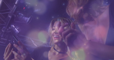 Two women wearing dark tops holding smartphone, leaning and smiling at club, truss and bokeh lights....