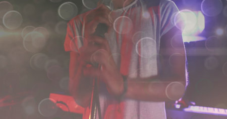 Gripping mic stand male singer performing on dark stage under spotlights, with towel and keyboard....