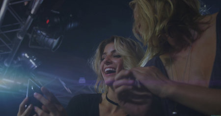 Smiling women checking smartphone screen at club stage, with metal truss and stage spotlights....