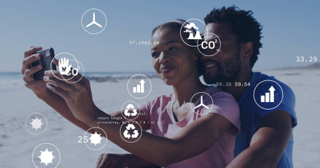 Couple taking selfie and sitting closely on sandy shoreline, with phone and floating eco data icons. Romantic, coastal, sustainability, urbanization, environmental, digital, sereneの写真素材