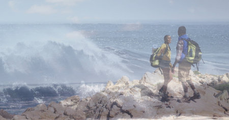 Holding hands couple standing on rocky coastal outcrop, with hiking backpacks and boots, copy space....