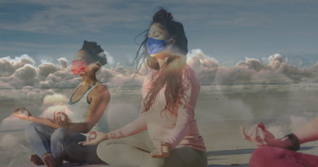 Meditating group of women cross-legged holding mudra on beach, wearing face masks and yoga mats....