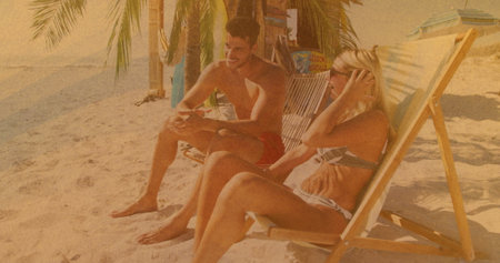 Sitting couple in swimwear holding smartphone, adjusting hair on chairs at palm-frond beach shelter. Tropical, relaxation, outdoor, leisure, vacation, sunshine, scenicの写真素材