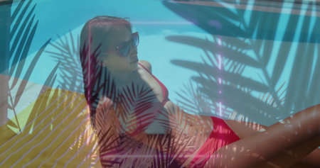 Lying Chinese woman wearing red bikini shades on lounger by turquoise pool, showing palm shadows....