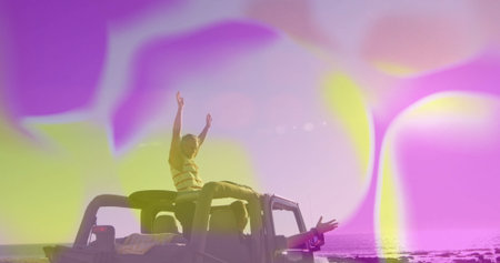Friends raising arms and leaning from open-top jeep on rocky shoreline, with colored bokeh overlays....