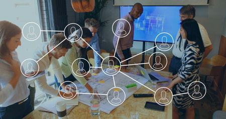 Collaborating business team of seven leaning over table in office, with plans and network overlay....