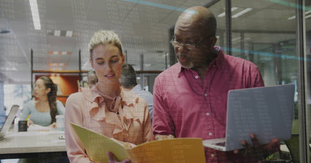 Collaborating senior man in maroon shirt and woman in peach blouse reviewing folder at office space....
