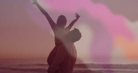Couple silhouette lifting woman overhead at beach shoreline during sunset, with ocean waves....