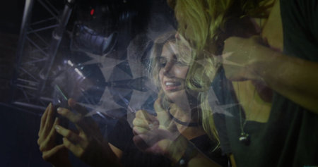 Holding smartphone, two friends leaning towards screen at concert, with geometric light overlay....