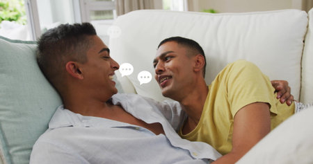 Reclining Indian men wearing casual tops at home on white sofa with speech bubbles near window. Modern, lounging, communication, intimacy, comfort, home decor, relaxedの写真素材