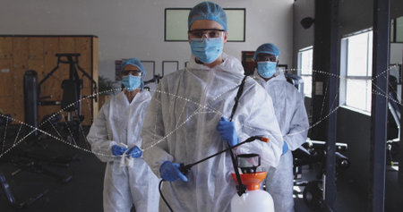 Disinfecting worker wearing white coveralls, hair nets, goggles and gloves at gym with sprayer wand....