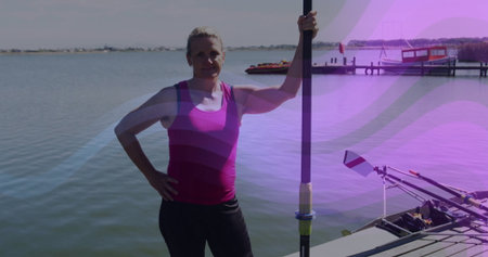 Standing woman wearing pink athletic top and black leggings on dock, holding rowing oar with shell....