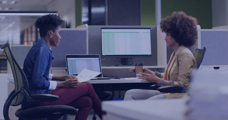 Holding reports and coffee cup, two colleagues reviewing spreadsheet data in open-plan office....