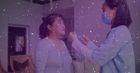 Offering scrubs-clad nurse holding surgical mask to seated patient in living room, with cushions....