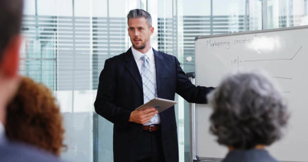 Presenting suited businessman holding tablet, gesturing to whiteboard in meeting room, with charts. Corporate, teamwork, professionalism, modern, communication, strategy, collaborationの写真素材