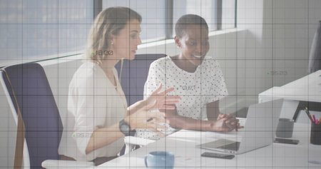 Two women wearing formal attire reviewing charts in office, with silver laptop and grid overlays. Collaboration, teamwork, professional, data analysis, modern, workspace, productivityの写真素材