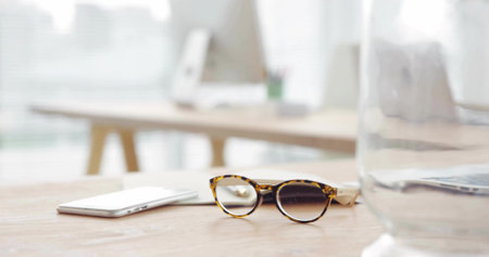 Featuring tortoiseshell glasses resting on wood desk in office, smartphone, laptop, water glass. Workspace, productivity, minimalism, technology, office accessories, modern, professionalの写真素材
