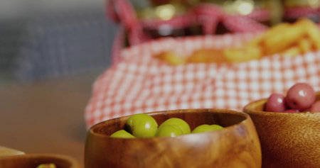 Displaying wooden bowl holding green olives on dining table, with purple olives and picnic basket. Rustic, mosaic, culinary, snack, arrangement, leisure, ambianceの写真素材