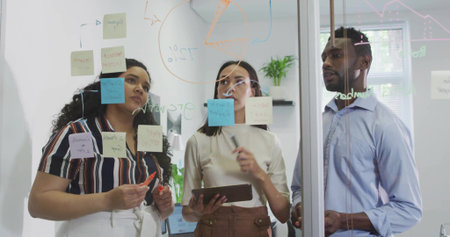 Brainstorming coworkers examining sticky notes on glass board in meeting room with tablet, markers....