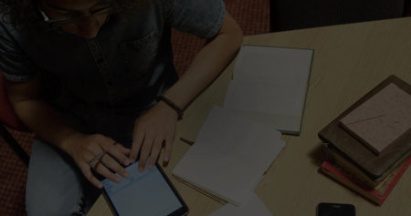 Tapping denim-clad student with glasses using touchscreen tablet at desk, with notebook, smartphone....