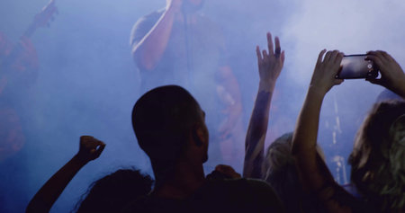 Silhouetted concertgoer holding smartphone watching performer gripping microphone at foggy hall....