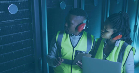 IT techs in vests earmuffs inspecting server racks in data center aisle, with laptop clipboard. Technology, infrastructure, cybersecurity, engineering, collaboration, modern, industrialの写真素材