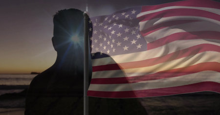Silhouetted man holding waving American flag on shore at sunset, with sunburst and ocean waves. Patriotism, freedom, determination, serenity, nature, silhouette, horizonの写真素材