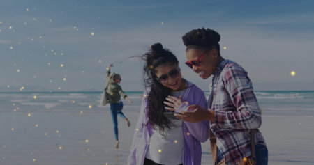 Leaping woman and friends leaning sharing smartphone on beach shoreline, with bags and sunglasses. Carefree, camaraderie, relaxation, lifestyle, outdoor, vibrant, leisureの写真素材