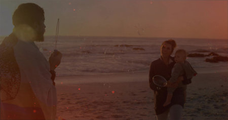 Man wearing towel, woman holding infant at sunset shoreline, blowing soap bubbles with bubble wand. Family, leisure, celebration, tranquility, harmony, outdoor, natural lightの写真素材