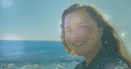 Smiling woman in patterned blouse standing at seaside with ocean waves and lens flare, copy space....