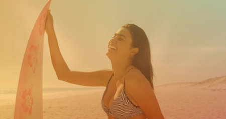 Smiling Asian woman in striped bikini holding pink floral surfboard on beach at sunset, copy space....