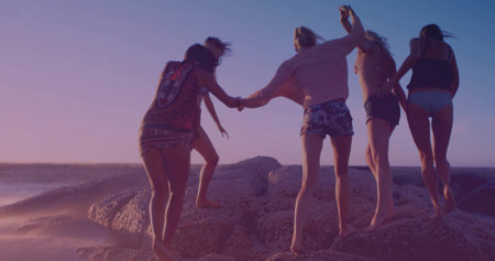 Walking five women in swimwear holding hands crossing barnacle-covered rocks at beach at sunset. Female camaraderie, coastal adventure, sunset glow, carefree lifestyle, nature connection, friendship travel, vibrant paletteの写真素材