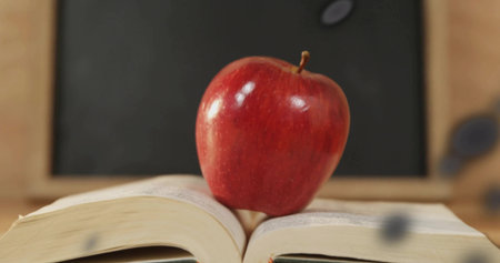 Sitting red apple balancing on open book on desk in classroom, with chalkboard and chalk dust. Stationery, education, learning, literary, vintage, academic, intellectualの写真素材