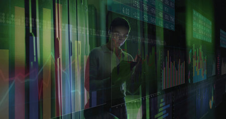 Standing white mid-adult woman wearing blouse holding tablet in tech workspace, with data panels....
