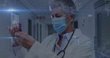 Inspecting senior doctor wearing lab coat and mask in hospital corridor holding blood tube. Medical, healthcare, clinical, research, diagnostics, supervision, professionalismの写真素材