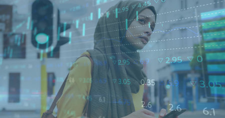 Standing woman in hijab holding smartphone at crossing traffic light, displaying financial overlay....